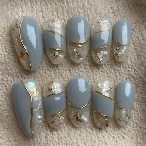 May include: Set of ten artificial nails in a light blue hue, adorned with gold accents and iridescent flakes. Each nail features a unique design with a combination of solid color, gold lines, and decorative elements. The nails are oval-shaped.
