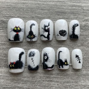 May include: A set of ten press-on nails with a white base and black cat illustrations. The designs feature various black cats with yellow eyes, a ball of yarn, and a fish skeleton. Each nail has a smooth, glossy finish and rounded edges.