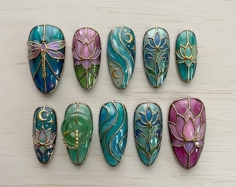 Peut inclure: Dix faux ongles aux motifs détaillés. Les ongles présentent des motifs tels qu'une libellule, des fleurs de lotus et des tourbillons d'eau, dans des tons de teal, de violet et d'or. Chaque ongle ovale a une finition brillante. Les motifs rappellent le vitrail.