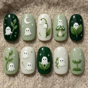 May include: A set of ten press-on nails featuring a palette of dark green, sage green, and white. The designs include raised 3D white ghosts, green leaves, white lily of the valley flowers, a crescent moon, stars, and a mushroom with a ghost.