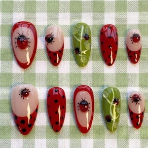 May include: A set of ten press-on nails featuring a ladybug theme. The designs include red and black polka dot patterns, clear sections with raised 3D ladybug embellishments, and green leaf motifs with small ladybug accents on a glossy finish.