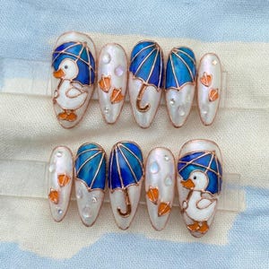 May include: A set of ten artificial nails with a white base, featuring designs of blue umbrellas, duck footprints, and a cartoon duck with an umbrella. The nails have a glossy finish and are adorned with small, clear embellishments.