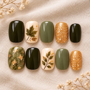 May include: A set of ten press-on nails featuring a variety of designs including solid dark forest green, muted olive green, cream with brown maple leaf illustrations, cream with green leaf sprigs, and gold glitter finishes with gold leaf accents.