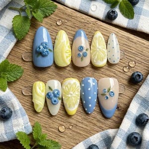 May include: A set of ten artificial nails with fruit-themed designs. The nails feature blueberries, lemon slices, and polka dots in shades of blue, yellow, and white. The nails are arranged on a wooden surface.