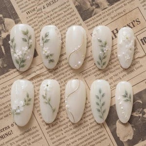 May include: A set of ten almond-shaped press-on nails in an off-white base color. The designs feature hand-painted green leaves, small white flowers, thin gold metallic lines, and tiny white pearl accents arranged in various floral patterns.