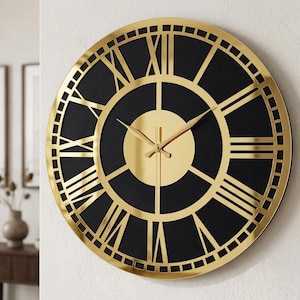 Puede incluir: Un reloj de pared grande y redondo con una esfera negra y números romanos y manecillas doradas. El reloj tiene un marco dorado y un diseño circular. El reloj está montado en una pared blanca. El diámetro del reloj es de aproximadamente 60 cm.