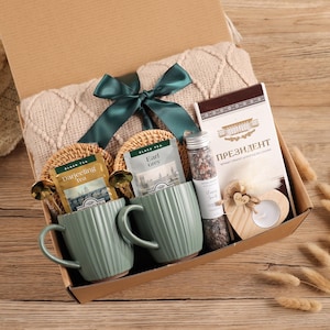 May include: A gift box containing tea, mugs, and a blanket. The box includes two green mugs, tea bags, a small jar, a chocolate bar, and a wooden heart-shaped candle holder. A green ribbon decorates the box, and a beige blanket is visible.