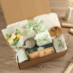 May include: A gift box containing a white blanket, a bouquet of yellow and white flowers, a candle, a floral mug, a tea box, and a small woven coaster. The box is tied with light green ribbons.