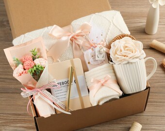 May include: A gift box filled with various items, including a white mug with a large rose, a notebook, a bouquet of flowers, socks, and a blanket. The items are in shades of white, pink, and beige, with satin ribbons. The box is brown.