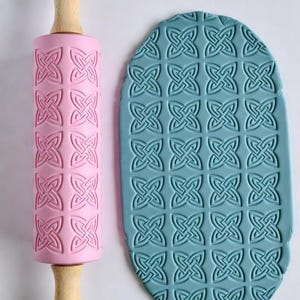 May include: A pink rolling pin with a Celtic knot pattern and wooden handles, next to a blue clay sheet with the same embossed design. The rolling pin is designed for creating textured patterns on dough or clay.