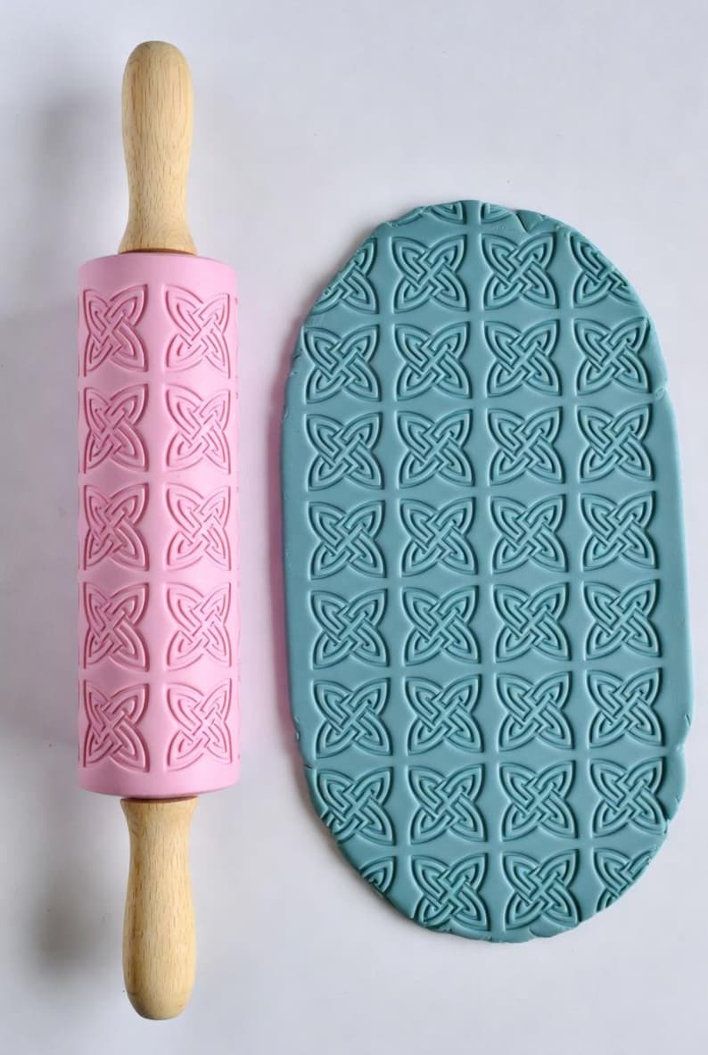 May include: A pink rolling pin with a Celtic knot pattern and wooden handles, next to a blue clay sheet with the same embossed design. The rolling pin is designed for creating textured patterns on dough or clay.