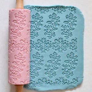 May include: A pink rolling pin with a snowflake pattern next to a piece of blue dough with the same snowflake design. The rolling pin has a wooden handle. The dough is imprinted with the snowflake pattern.