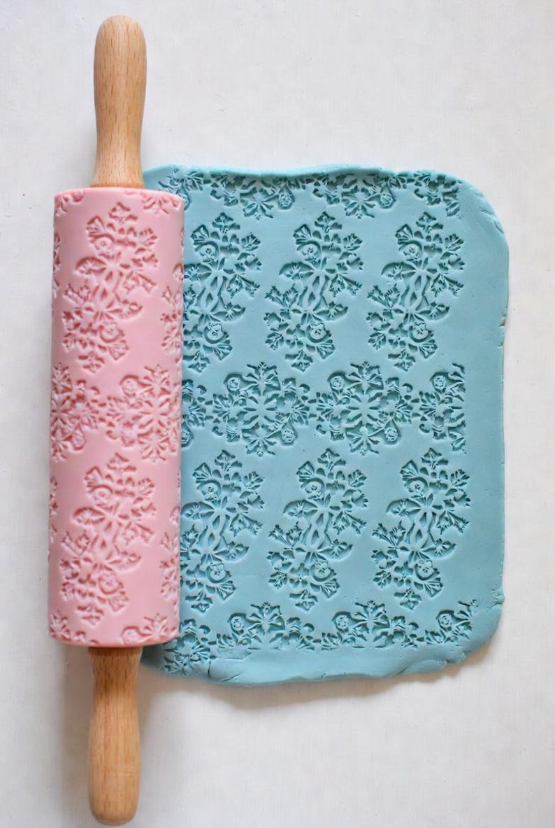 May include: A pink rolling pin with a snowflake pattern next to a piece of blue dough with the same snowflake design. The rolling pin has a wooden handle. The dough is imprinted with the snowflake pattern.
