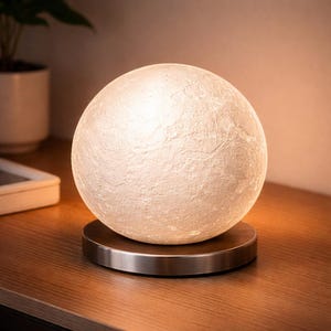 May include: A spherical, textured lamp with a bright, warm glow, resembling the moon. The lamp sits on a circular, brushed metal base. The lamp is a decorative lighting fixture.