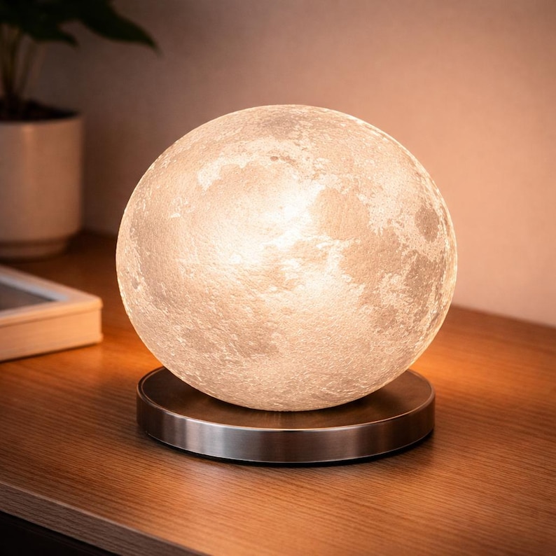 May include: A spherical, illuminated moon lamp with a textured surface, resting on a circular, brushed metal base. The lamp emits a soft, warm glow, creating a celestial ambiance. The lamp is a decorative lighting fixture.