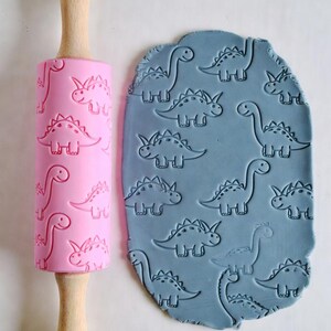 May include: A pink rolling pin with a wooden handle and a dinosaur pattern next to a piece of blue clay imprinted with the same dinosaur design. The rolling pin is made of silicone and wood, and the clay is a light blue color.