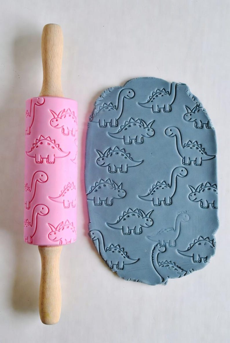 May include: A pink rolling pin with a wooden handle and a dinosaur pattern next to a piece of blue clay imprinted with the same dinosaur design. The rolling pin is made of silicone and wood, and the clay is a light blue color.
