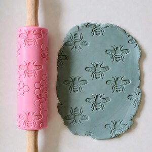 May include: A pink rolling pin with a bee and honeycomb pattern next to a piece of blue clay imprinted with the same bee design. The rolling pin has wooden handles. The bee design is repeated across the rolling pin and clay.