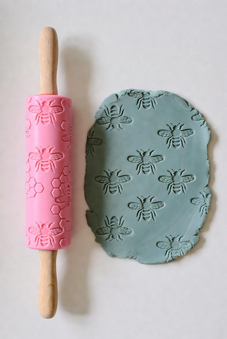 May include: A pink rolling pin with a bee and honeycomb pattern next to a piece of blue clay imprinted with the same bee design. The rolling pin has wooden handles. The bee design is repeated across the rolling pin and clay.