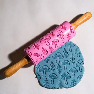 May include: A pink rolling pin with a mushroom and leaf pattern rests on a blue, textured dough. The rolling pin has wooden handles. The dough is imprinted with the same mushroom and leaf design.