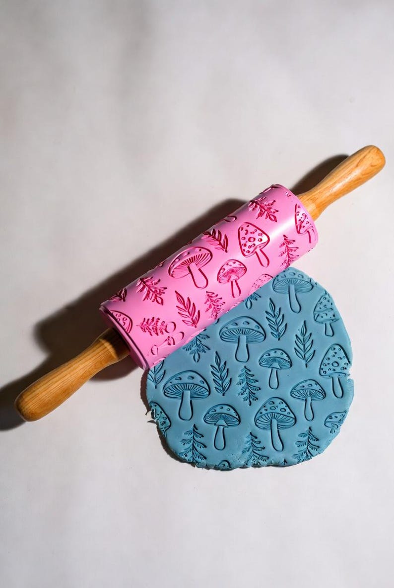 May include: A pink rolling pin with a mushroom and leaf pattern rests on a blue, textured dough. The rolling pin has wooden handles. The dough is imprinted with the same mushroom and leaf design.