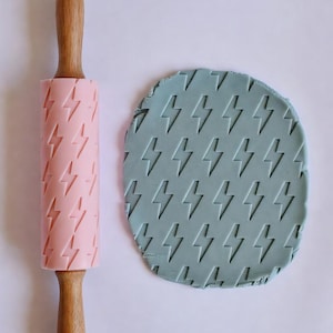May include: A pink rolling pin with wooden handles and a lightning bolt pattern next to a circle of blue-green dough with the same lightning bolt design. The rolling pin is for baking.