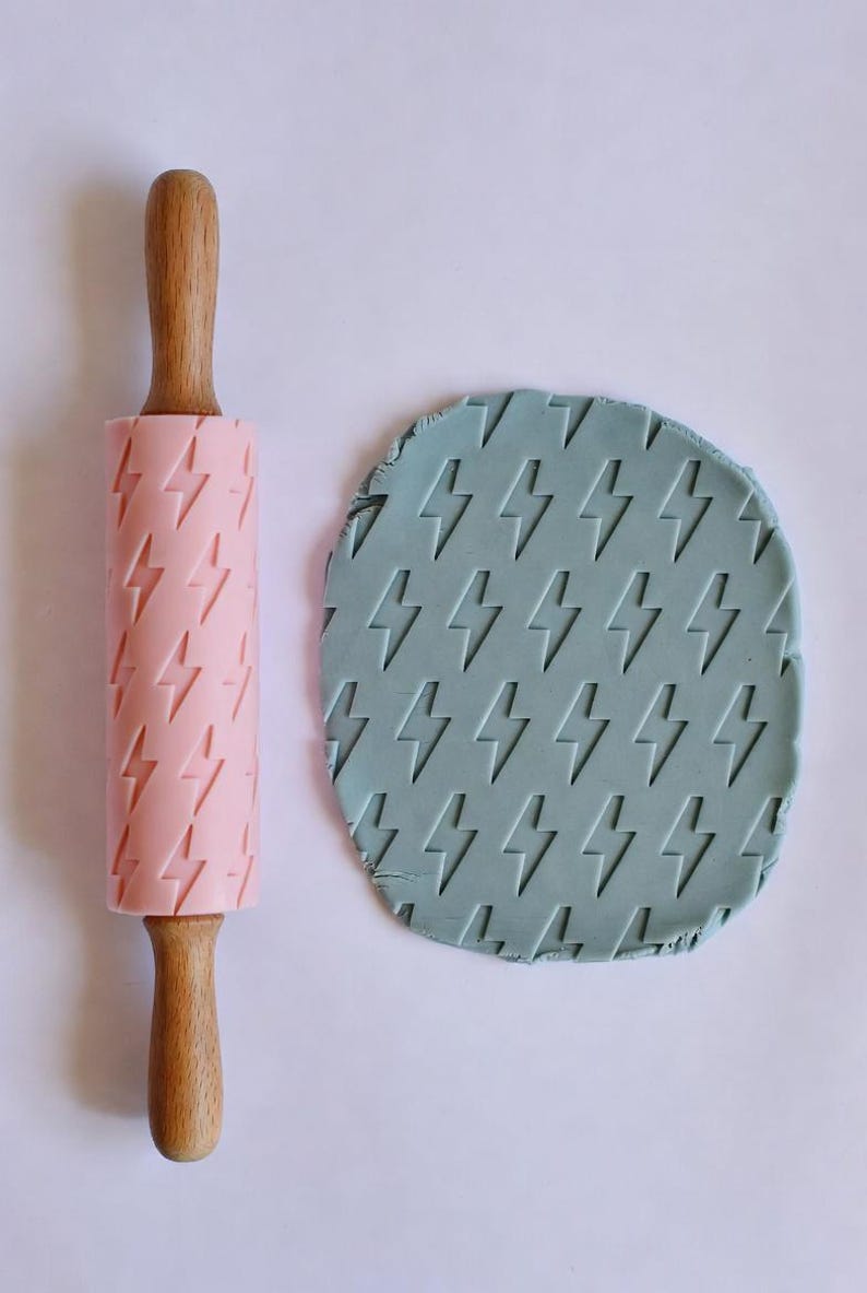 May include: A pink rolling pin with wooden handles and a lightning bolt pattern next to a circle of blue-green dough with the same lightning bolt design. The rolling pin is for baking.