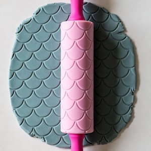 May include: A pink rolling pin with a mermaid scale pattern, resting on a piece of gray clay with the same pattern. The rolling pin has a wooden handle and pink grips.