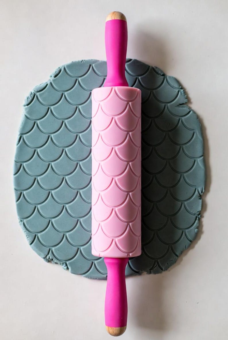 May include: A pink rolling pin with a mermaid scale pattern, resting on a piece of gray clay with the same pattern. The rolling pin has a wooden handle and pink grips.