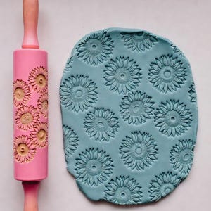 May include: A pink rolling pin with a sunflower pattern next to a piece of blue clay imprinted with the same sunflower design. The rolling pin has wooden handles. The clay is oval-shaped.