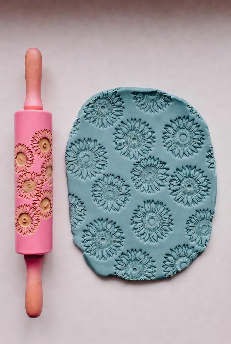 May include: A pink rolling pin with a sunflower pattern next to a piece of blue clay imprinted with the same sunflower design. The rolling pin has wooden handles. The clay is oval-shaped.