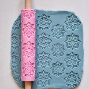 May include: A pink rolling pin with a floral pattern is pressed into a sheet of blue clay, creating an embossed design. The rolling pin has a wooden handle. The clay is covered in a repeating floral pattern.