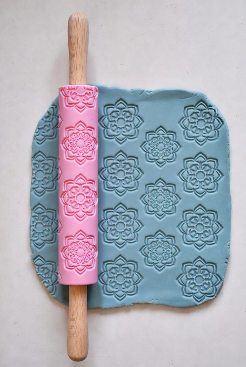 May include: A pink rolling pin with a floral pattern is pressed into a sheet of blue clay, creating an embossed design. The rolling pin has a wooden handle. The clay is covered in a repeating floral pattern.