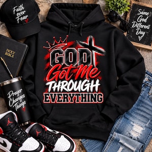May include: A black hooded sweatshirt featuring a graphic with a red crown, a black cross, and the white and red text "GOD Got Me THROUGH EVERYTHING". The hoodie is displayed alongside a black baseball cap, a black tumbler, a Bible, and sneakers.