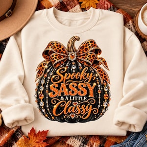 Op de afbeelding: Een crèmekleurige sweater met ronde hals en een afbeelding van een zwarte pompoen versierd met een strik met luipaardprint en fonkelende strass-steentjes. De tekst luidt Spooky, SASSY & A LITTLE Classy in oranje en gouden letters.