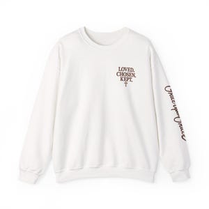 May include: A white crewneck sweatshirt featuring brown text on the upper left chest that reads LOVED. CHOSEN. KEPT. with a small cross symbol below it. The left sleeve features the brown cursive text Grace upon Grace running vertically.