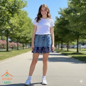 May include: A denim skirt with a blue paisley patterned ruffle. The skirt is paired with a white t-shirt and white sneakers. The image is taken outdoors on a sunny day.