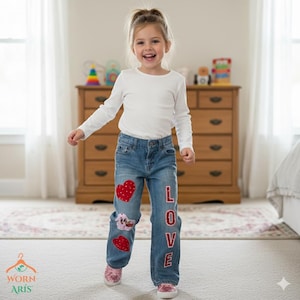 May include: A young girl wearing blue denim jeans with red heart patches and the word "LOVE" appliqued down the side. She is also wearing a white long-sleeve shirt and pink shoes. The background includes a wooden dresser and a window.