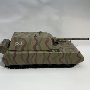 May include: A model tank with a camouflage pattern in shades of brown, green, and tan. The tank has a long barrel and the number "S33" is visible on the side. The tank is a military collectible.