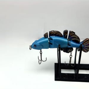 May include: A blue and brown articulated fishing lure shaped like a fish. The lure has a segmented body, two treble hooks, and a detailed paint job. It is mounted on a black stand, showcasing its design and features.