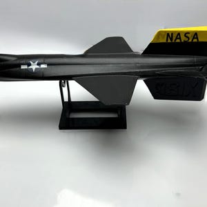 Pode incluir: Um modelo de avião preto com asas e cauda cinzentas, e uma barbatana traseira amarela e preta com "NASA" impressa. O modelo tem um emblema de estrela branca e é montado num suporte preto.