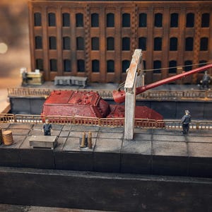 Puede incluir: Diorama en miniatura de una escena de astillero con una grúa roja, un tanque rojo y figuras en miniatura. El telón de fondo es un edificio de ladrillo. Hay barriles y otros detalles.