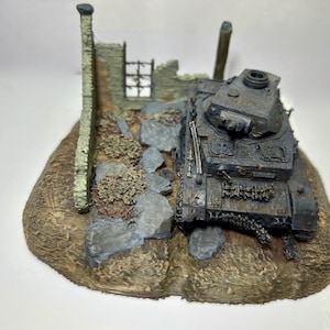 May include: A detailed miniature model of a dark gray tank, positioned on a diorama base. The base features a textured, brown terrain with gray rocks, and a weathered, stone wall. The tank is a military vehicle.