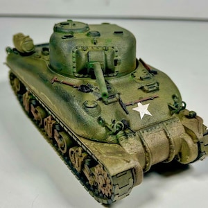 Op de afbeelding: Een schaalmodel van een olijfgroene Sherman-tank met een draaibare koepel, een centraal kanon en een wit sterembleem op de voorzijde. De tank heeft gedetailleerde rupsbanden, wielen en kleine gereedschappen bevestigd aan de buitenkant van het voertuig.