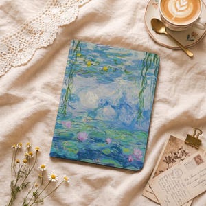 May include: A notebook featuring a Monet-style water lilies painting in shades of blue, green, and pink. The notebook is lying on a cream-colored surface, with a cup of coffee and postcards nearby.