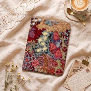 Puede incluir: Un cuaderno rectangular con un patrón de puntos intrincado y vibrante en tonos rojo, amarillo, negro y azul. El diseño incluye motivos circulares, formas de estrella y líneas fluidas que cubren toda la superficie de la portada.