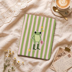 May include: A rectangular tablet case featuring a vertical striped pattern in light green and pale pink. Centered on the case is an illustration of a green frog wearing dark green boots with heart details and holding a pink hat, surrounded by yellow stars.