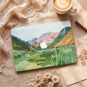Op de afbeelding: Een laptophoes met een landschapsschilderij van bergen, een meer en groene velden. De hoes heeft een wit Apple-logo. De scène is geschilderd in tinten groen, blauw, oranje en bruin. Een kopje koffie, een boek en een ansichtkaart staan ook op de foto.
