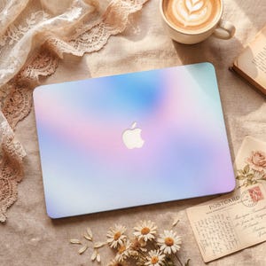 Może przedstawiać: Laptop z pastelowym gradientem w odcieniach niebieskiego, różowego i fioletowego. Logo Apple jest widoczne na powierzchni laptopa. Laptop leży na beżowej powierzchni z filiżanką kawy, książką i pocztówką.