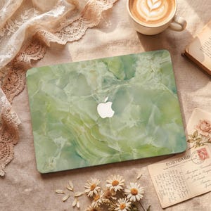 Può includere: Una cover per laptop verde con motivo marmorizzato e logo Apple bianco. La cover è su una superficie beige, con una tazza di caffè, una cartolina, un libro e margherite. La cover ha un aspetto marmorizzato in pietra naturale.